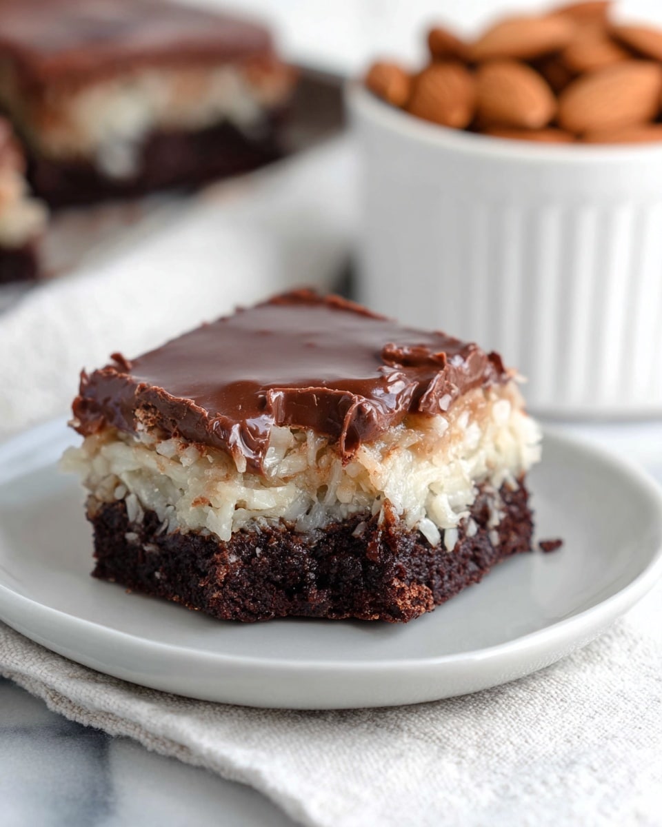 A close-up of a two-layer square dessert on a simple white plate, placed on a white marbled surface with a white and gray cloth underneath. The bottom layer is thick, dark brown, and dense, showing a rich chocolate cake base. The middle layer is lighter in color, creamy with a rough texture from shredded coconut that looks moist and soft. The top layer is a smooth, glossy chocolate glaze with a few small imperfections and a slightly uneven surface. In the blurred background, there is a white ceramic container filled with whole almonds, adding a warm and natural touch to the scene. photo taken with an iphone --ar 4:5 --v 7