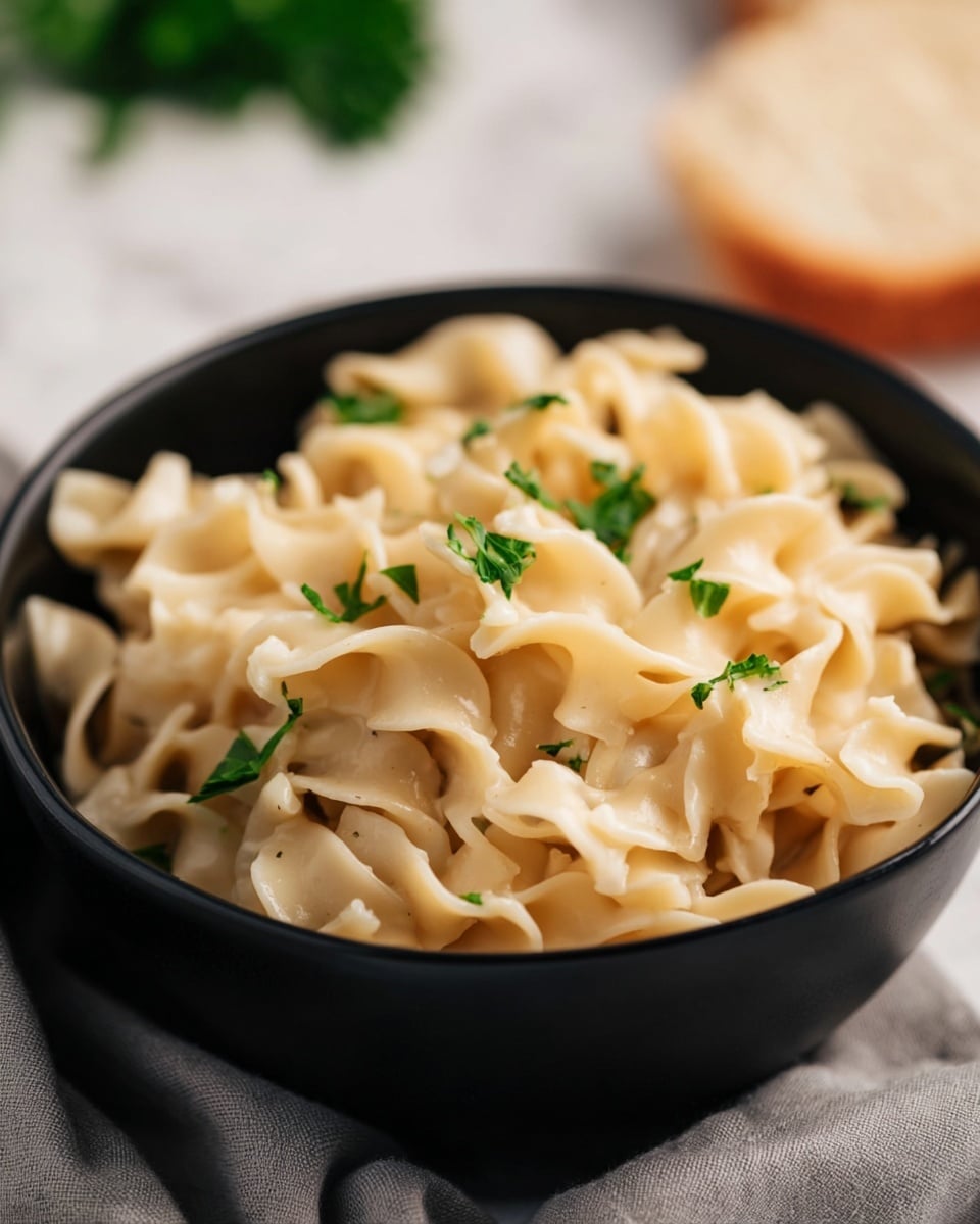 A close-up of a black bowl filled with soft, creamy light beige pasta noodles that have a wavy texture. Small green parsley leaves are scattered on top, adding a pop of color. The bowl sits on a light gray cloth, placed on a surface with a white marbled texture. In the soft background, there is a blurred piece of bread and some green herbs. Photo taken with an iphone --ar 4:5 --v 7