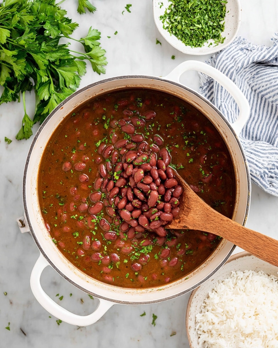 A white pot filled with dark red beans cooked in a rich brown broth, topped with small green parsley bits scattered across the surface; a wooden spoon rests inside the pot, scooping up a pile of beans and herbs. The pot is placed on a white marbled surface, with fresh green parsley on the top left, a white small dish of chopped parsley to the bottom left, and a white bowl of plain white rice to the bottom right, partly visible. A white and blue striped cloth is placed at the top right corner. Photo taken with an iphone --ar 4:5 --v 7