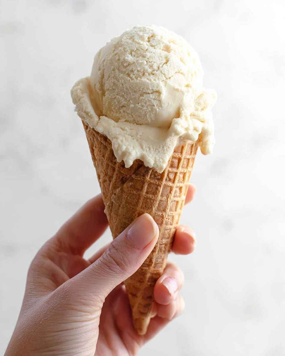 A close-up view of a single scoop of creamy, light beige ice cream with a smooth, slightly rough texture, sitting on top of a medium brown waffle cone with a grid pattern. The ice cream scoop is rounded with soft edges, slightly overflowing the cone. A woman's hand is holding the cone from the bottom, showing natural skin tone and short nails. The background is bright and plain with a white marbled texture. Photo taken with an iphone --ar 4:5 --v 7
