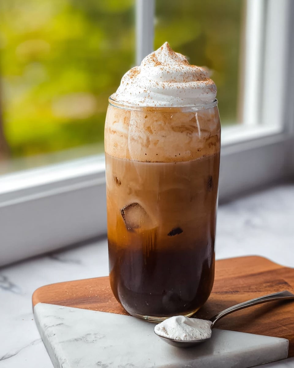 A tall clear glass filled with three layers: the bottom layer is dark brown iced coffee with visible ice cubes, the middle layer is a thick light brown foam, and the top layer is a swirl of white whipped cream sprinkled with light brown powder. The glass sits on a wooden board placed on a white marbled surface. A spoon with some foam on it lies next to the glass. The background shows a window with green blurred trees outside. Photo taken with an iphone --ar 4:5 --v 7
