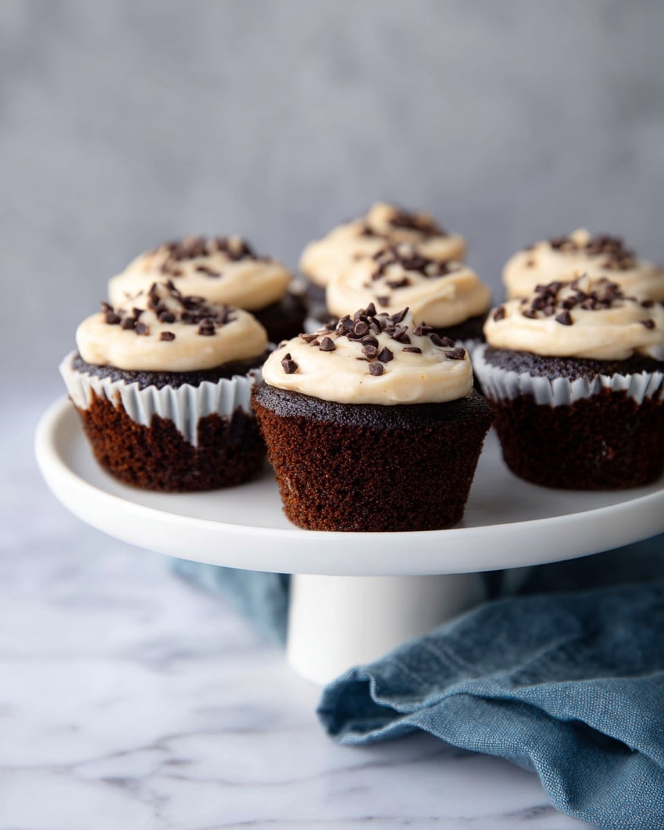 Six dark brown chocolate cupcakes are placed on a white cake stand with a white marbled background. Each cupcake has one thick layer of smooth, light cream-colored frosting on the top, sprinkled with small dark brown chocolate chips. The cupcakes are evenly spaced and the texture of the cake looks soft and moist. A blue cloth is casually placed under the cake stand. Photo taken with an iphone --ar 4:5 --v 7