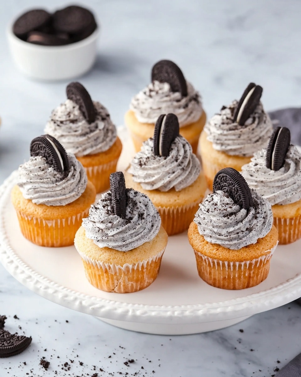 Seven vanilla cupcakes are arranged on a white plate with detailed edges, each topped with a generous swirl of light gray cookies and cream frosting speckled with cookie crumbs. A half Oreo cookie is placed upright on top of the frosting on each cupcake, adding a dark black contrast to the pale frosting and golden brown cupcakes. The white marbled surface beneath the plate complements the soft, light colors of the cupcakes. The background shows a small white bowl filled with more Oreo cookies and cookie crumbs scattered nearby. Photo taken with an iphone --ar 4:5 --v 7