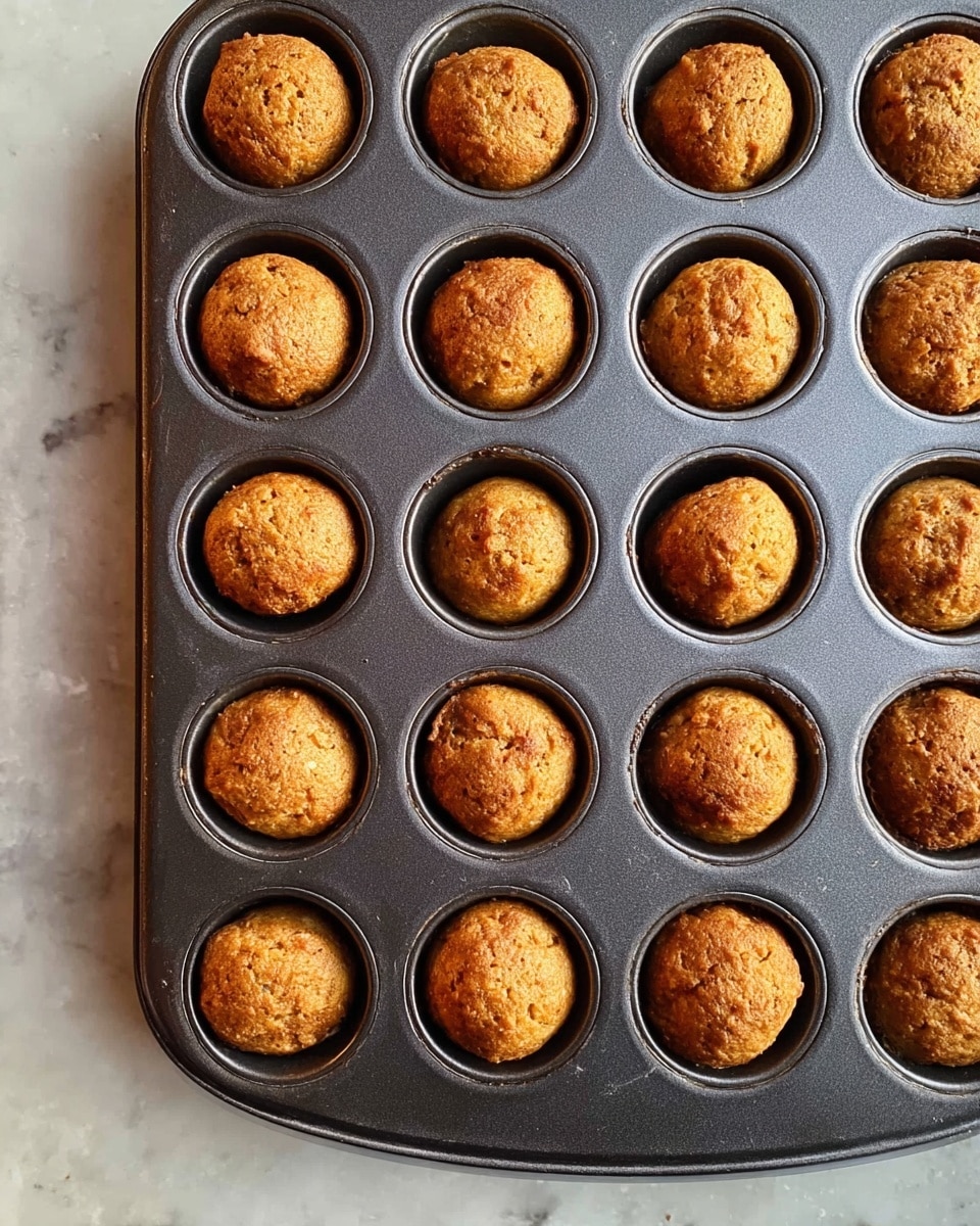 A dark metal mini muffin pan holds 24 small, round baked balls with a golden-brown color and a slightly rough texture. Each ball fills one cup of the tray, showing a lightly crisped surface with small uneven lumps. The pan rests on a white marbled surface, creating a clean contrast with the warm baked treats. Photo taken with an iphone --ar 4:5 --v 7