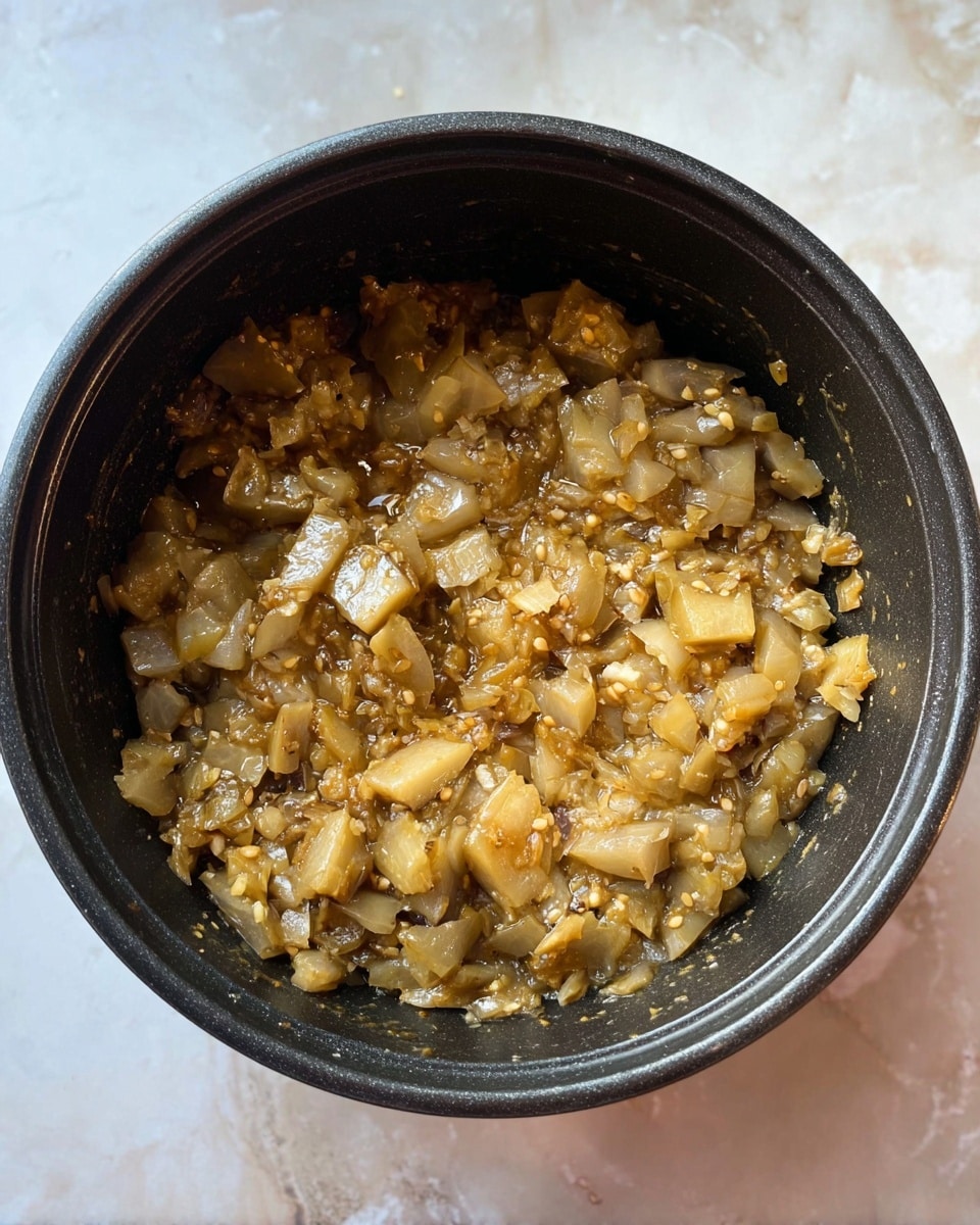 Inside a black cooking pot on a white marbled surface, there is a single layer of cooked eggplant pieces that are soft and moist. The eggplant pieces vary in size and shape, showing a mix of light brown skin and pale, translucent flesh with tiny white seeds visible throughout. The texture appears tender and slightly saucy, with some small bits resembling seeds spread evenly in the mixture. The pot's simple round shape frames the dish, which fills most of the bottom, highlighting the warm, earthy tones of the cooked eggplant. photo taken with an iphone --ar 4:5 --v 7