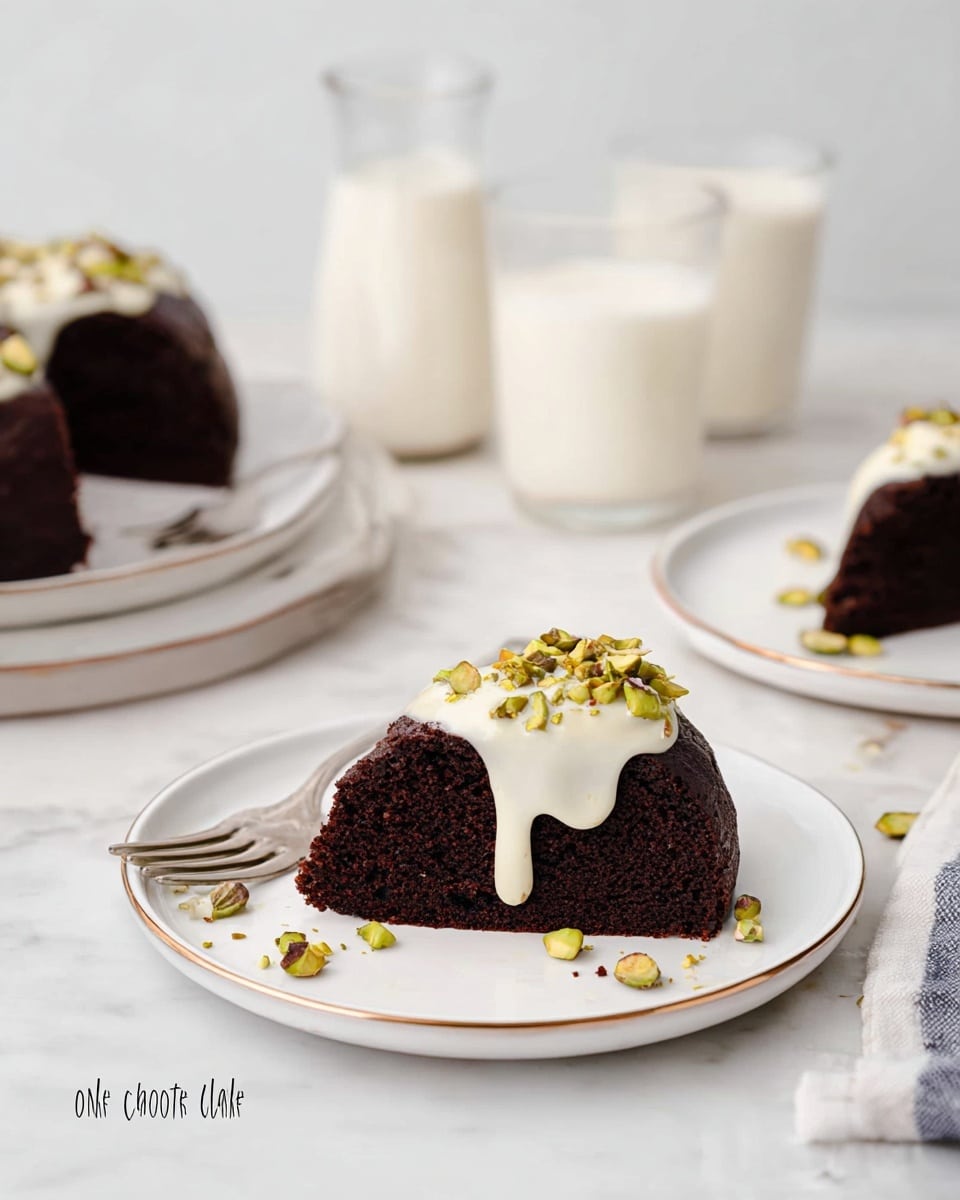The image shows a dark brown chocolate cake slice on a white plate with a thin gold rim. The cake has one layer with a smooth texture, topped with a creamy white sauce that drips slightly down the sides and is sprinkled with chopped green pistachios. In the background, there are two more white plates with similar cake slices and two clear glasses with milk, all placed on a white marbled surface. A silver fork is placed on the surface near the plate in the foreground. photo taken with an iphone --ar 4:5 --v 7