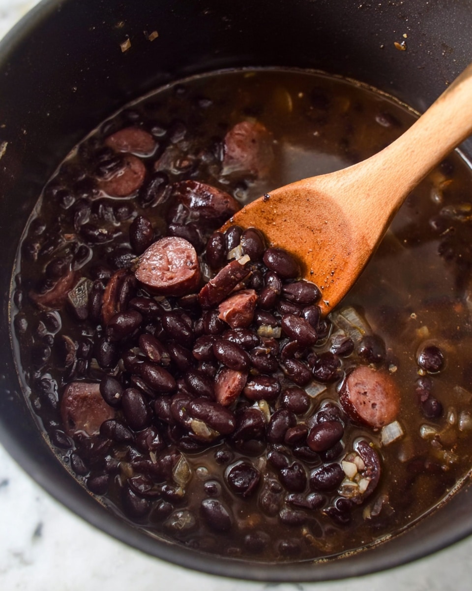 The image shows a pot of cooked black beans with sliced dark sausage pieces mixed in, and small bits of onion visible throughout the dish. The black beans are soft and glossy, floating in a thin dark brown broth. A wooden spoon is scooping up some of the beans and sausage, partially submerged in the bean mixture. The sides of the pot are dark and visible around the food. The scene is set on a white marbled surface. photo taken with an iphone --ar 4:5 --v 7