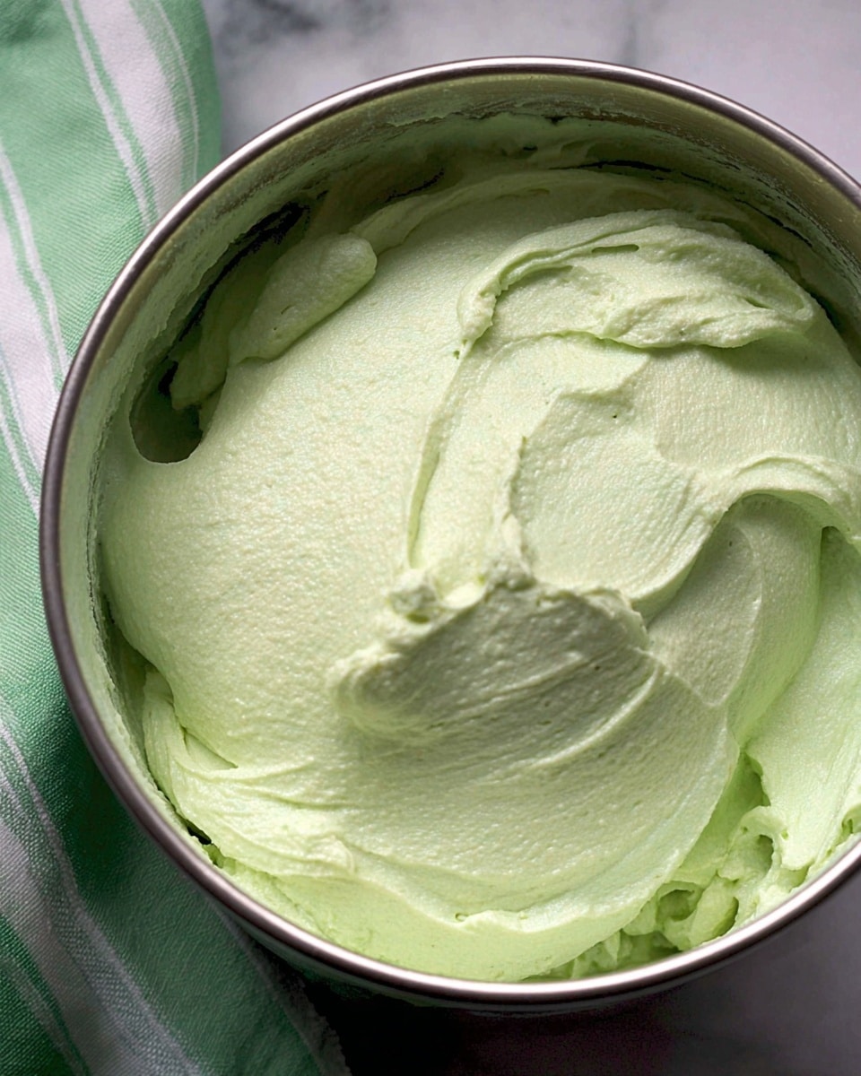 The image shows a close-up of a metal bowl filled with thick, light green creamy mixture with a smooth, whipped texture and soft peaks on the surface. The bowl sits on a white marbled texture, and there is a green and white striped cloth partially visible on the left side. Photo taken with an iphone --ar 4:5 --v 7