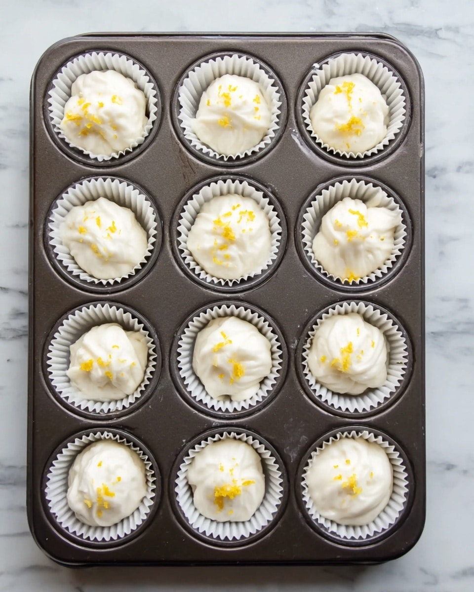 A dark metal baking tray with twelve square wells each lined with a white paper cup, filled with white creamy batter that is softly swirled on top; six of the batter cups have small sprinkles of yellow zest topping them, evenly spread across the tray on a white marbled surface photo taken with an iphone --ar 4:5 --v 7