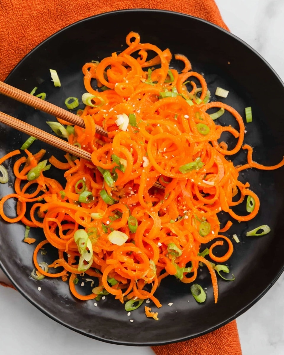A black round plate holds a single layer of bright orange curly carrot spirals spread out evenly across the surface. Scattered on top are small pieces of fresh green onion, adding light green and white pops of color. A pair of wooden chopsticks is lifting some carrot spirals from the left side of the plate. The plate is placed on a white marbled surface with an orange fabric in the background. photo taken with an iphone --ar 4:5 --v 7