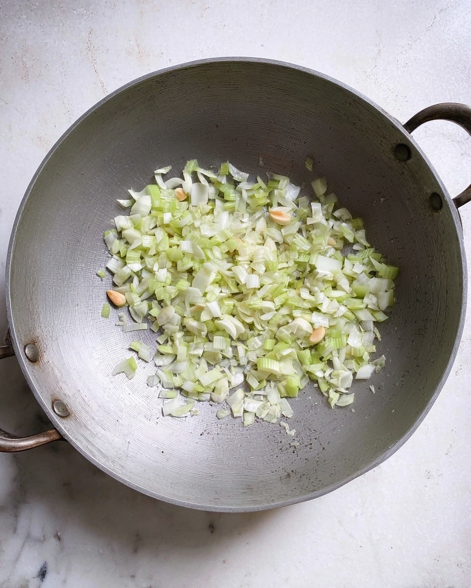 A large, round silver pan with two small handles contains finely chopped light green celery, white onion pieces, and small bits of pale garlic scattered in the center. The pan shows many light scratches from use. The background is a white marbled texture. photo taken with an iphone --ar 4:5 --v 7