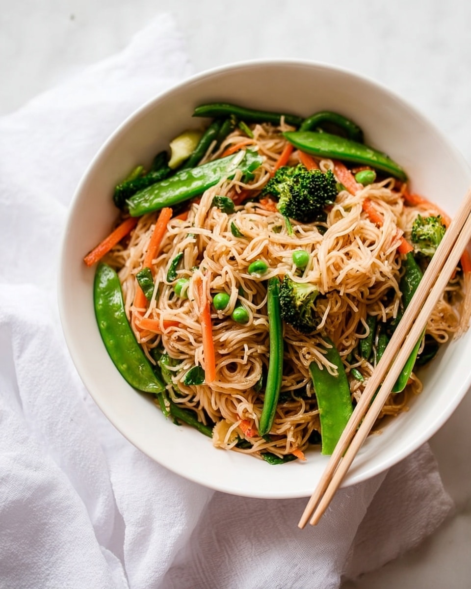 The image shows a white bowl filled with stir-fried noodles and vegetables placed on a white marbled surface. The noodles are light brown and sit in the middle layer, surrounded by bright green long beans, snap peas, and broccoli florets, with thin orange carrot strips scattered evenly throughout. A pair of light-colored chopsticks rests on the right side of the bowl, angled slightly down into the noodles. A soft white cloth is casually placed to the left side outside the bowl. The overall look is fresh and colorful with a focus on the mix of textures and vibrant vegetable colors. photo taken with an iphone --ar 4:5 --v 7