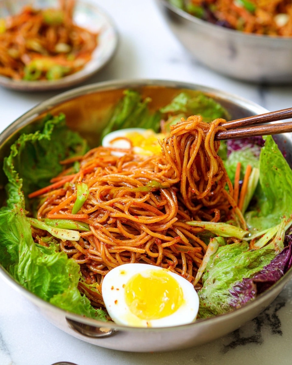 A close-up view of a silver bowl filled with a serving of spicy red noodles mixed with thin strips of vegetables such as carrots and cabbage, with fresh green leafy lettuce surrounding the noodles. A halved boiled egg with a bright yellow yolk rests on the side inside the bowl. Wooden chopsticks are lifting a bundle of noodles from the center of the bowl. The bowl sits on a white marbled surface, with a slightly blurred background showing another bowl and small plate. Photo taken with an iphone --ar 4:5 --v 7