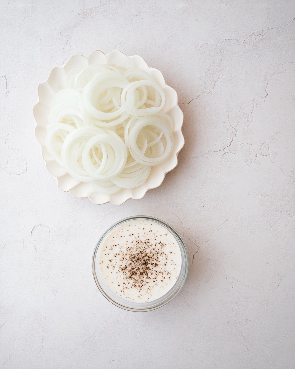 A white scalloped plate holds many thin, round, translucent white onion rings layered loosely on top of each other, creating soft, overlapping circles. Below it, a clear round bowl contains a creamy white sauce sprinkled with coarse black pepper, adding tiny dark specks on the smooth surface. Both dishes sit on a white marbled textured background with soft lighting, showing gentle shadows and cracks in the surface. photo taken with an iphone --ar 4:5 --v 7