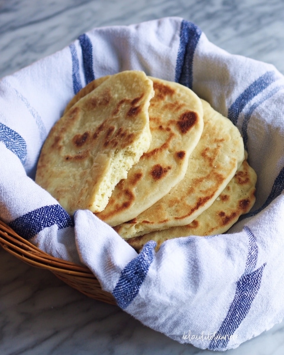 A basket lined with a white cloth with blue stripes holds four round flatbreads stacked on each other. The flatbreads have a light golden brown color with slightly darker patches that show they are cooked. One flatbread is broken in half, showing a soft and spongy texture inside. The basket sits on a white marbled surface. photo taken with an iphone --ar 4:5 --v 7
