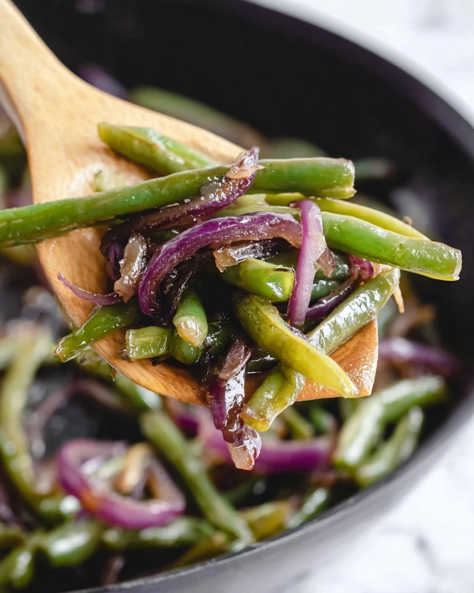 A close-up image showing a wooden spoon holding a mix of cooked vegetables, including green beans and thin slices of purple onion. The green beans are bright green and shiny, some with a slight yellowish tint, indicating they are cooked. The purple onions are soft, glossy, and appear slightly caramelized. The background is a black pan with more of the vegetable mix, placed on a white marbled surface. The focus is on the food on the spoon, giving a fresh and warm feeling. Photo taken with an iphone --ar 4:5 --v 7