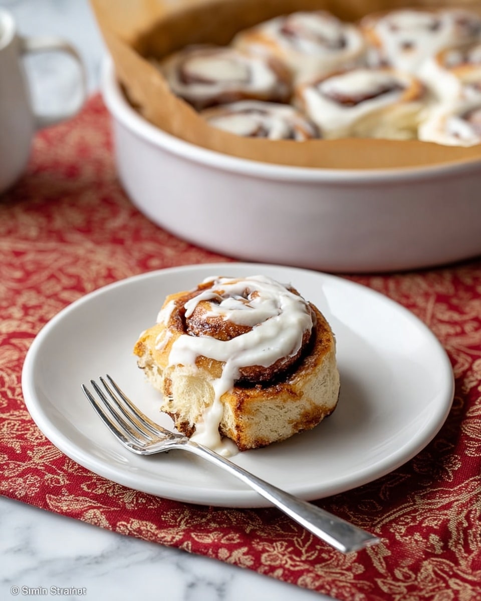 A round white bowl lined with light brown parchment paper holds eight cinnamon roll spirals arranged close together, each roll showing a golden-brown baked crust with darker brown cinnamon swirls visible in the center. The rolls have a soft, fluffy texture with some parts slightly crisped on top. The bowl sits on a white marbled surface covered partially by a red and white patterned cloth. The background has a warm wooden wall texture. photo taken with an iphone --ar 4:5 --v 7