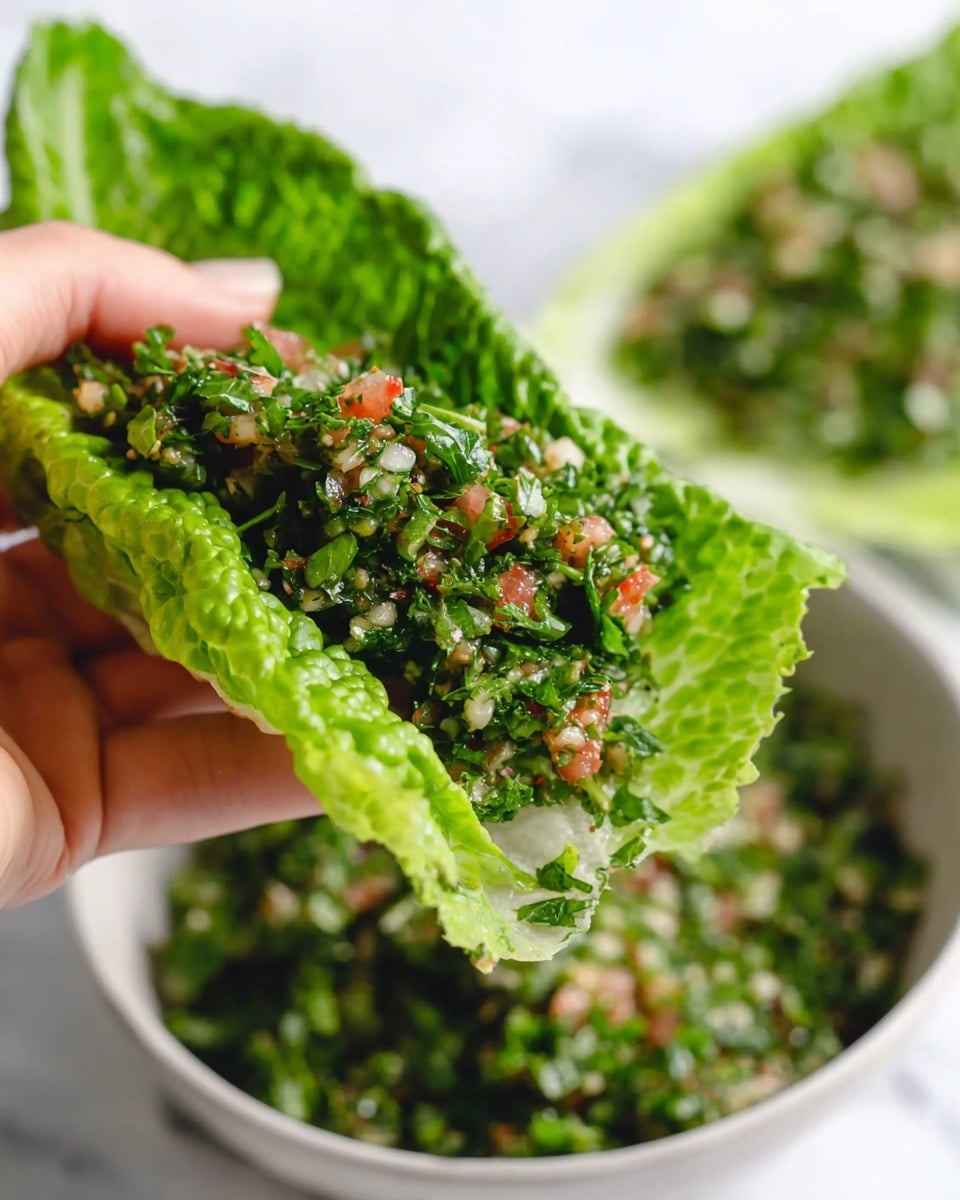 The image shows a woman's hand holding a bright green romaine lettuce leaf filled with a mix of finely chopped herbs and small pieces of tomato. The filling is rich green with specks of red and white, giving it a fresh and textured look. In the background, there is a white bowl full of the same herb mixture sitting on a white marbled surface that is softly blurred to keep focus on the lettuce leaf. The overall scene feels fresh and healthy with natural colors and clear details. photo taken with an iphone --ar 4:5 --v 7