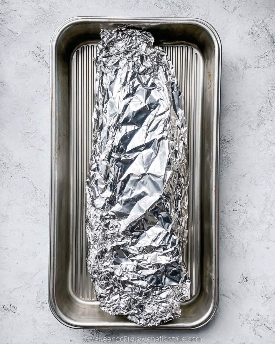 A long piece of food is fully wrapped in crumpled shiny silver foil placed in the middle of a metal roasting pan with ridges inside, the pan itself resting on a white marbled textured surface. photo taken with an iphone --ar 4:5 --v 7