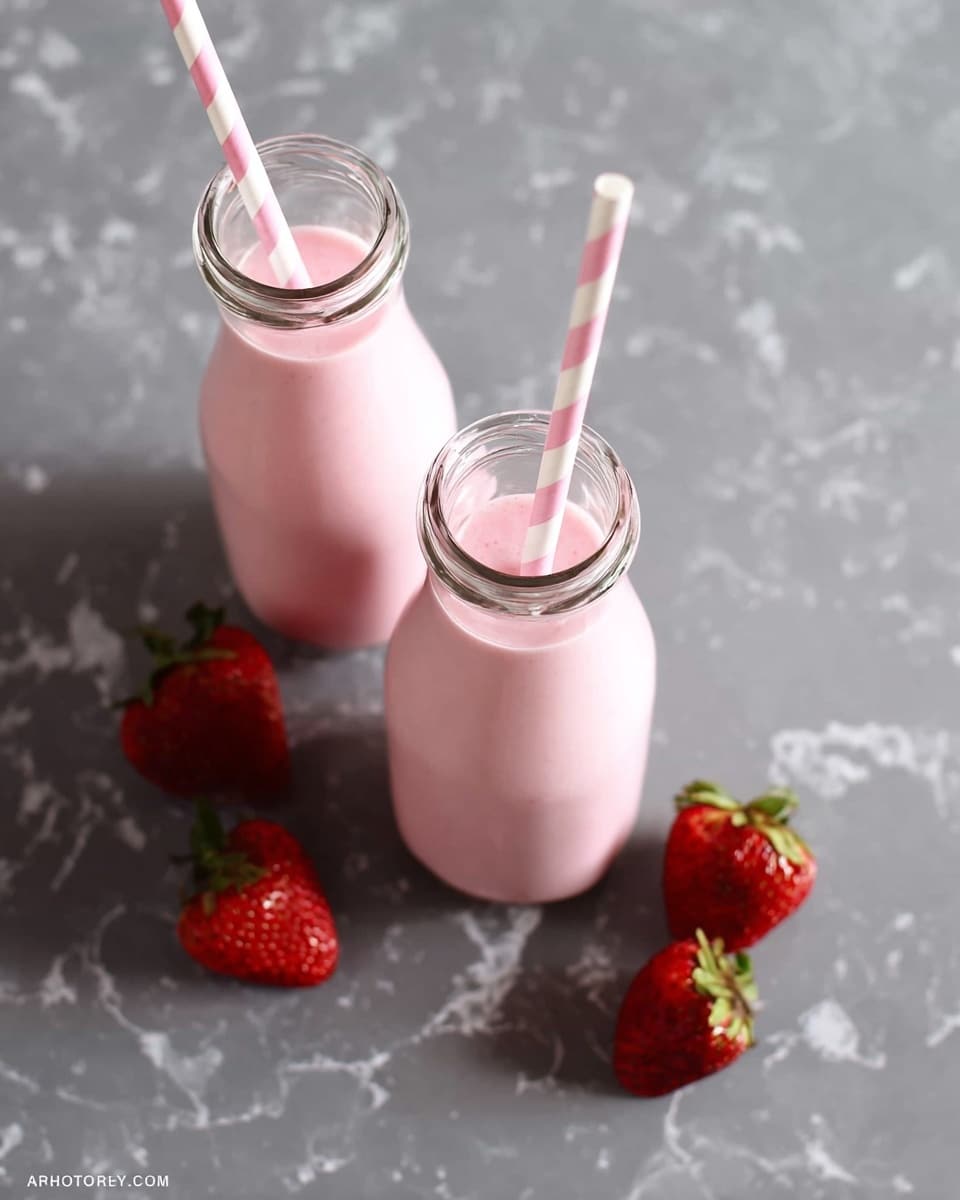 Two glass bottles filled with light pink strawberry smoothie, each with a white and pink striped straw, are placed on a white marbled surface. Around the bottles, there are five red strawberries with green tops scattered naturally. The scene shows a clean, fresh look with the smoothie texture smooth and creamy inside the clear bottles. Photo taken with an iphone --ar 4:5 --v 7