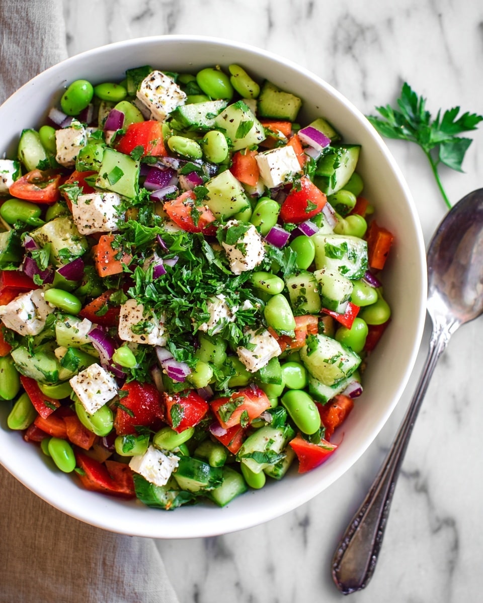 A white bowl filled with a colorful salad sits on a white marbled surface beside a silver spoon. The salad has four main layers: bright green broad beans form a thick base, mixed with chopped cucumber pieces showing light and dark green skin, and red bell pepper chunks adding a bright red contrast. Scattered among these are white cubes of feta cheese with a slightly crumbly texture and small pieces of diced purple onion. The salad is topped with finely chopped fresh parsley and a light sprinkling of black pepper. Photo taken with an iphone --ar 4:5 --v 7