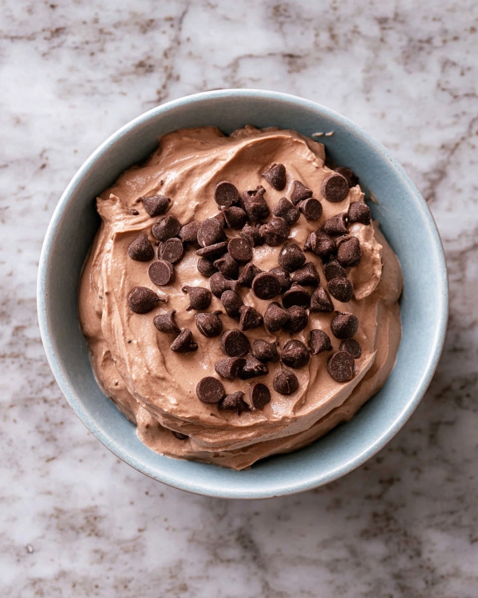 A light blue bowl filled with one layer of smooth, creamy chocolate mousse in a soft brown color, topped evenly with scattered dark brown chocolate chips on top. The bowl is placed on a white marbled textured surface. photo taken with an iphone --ar 4:5 --v 7
