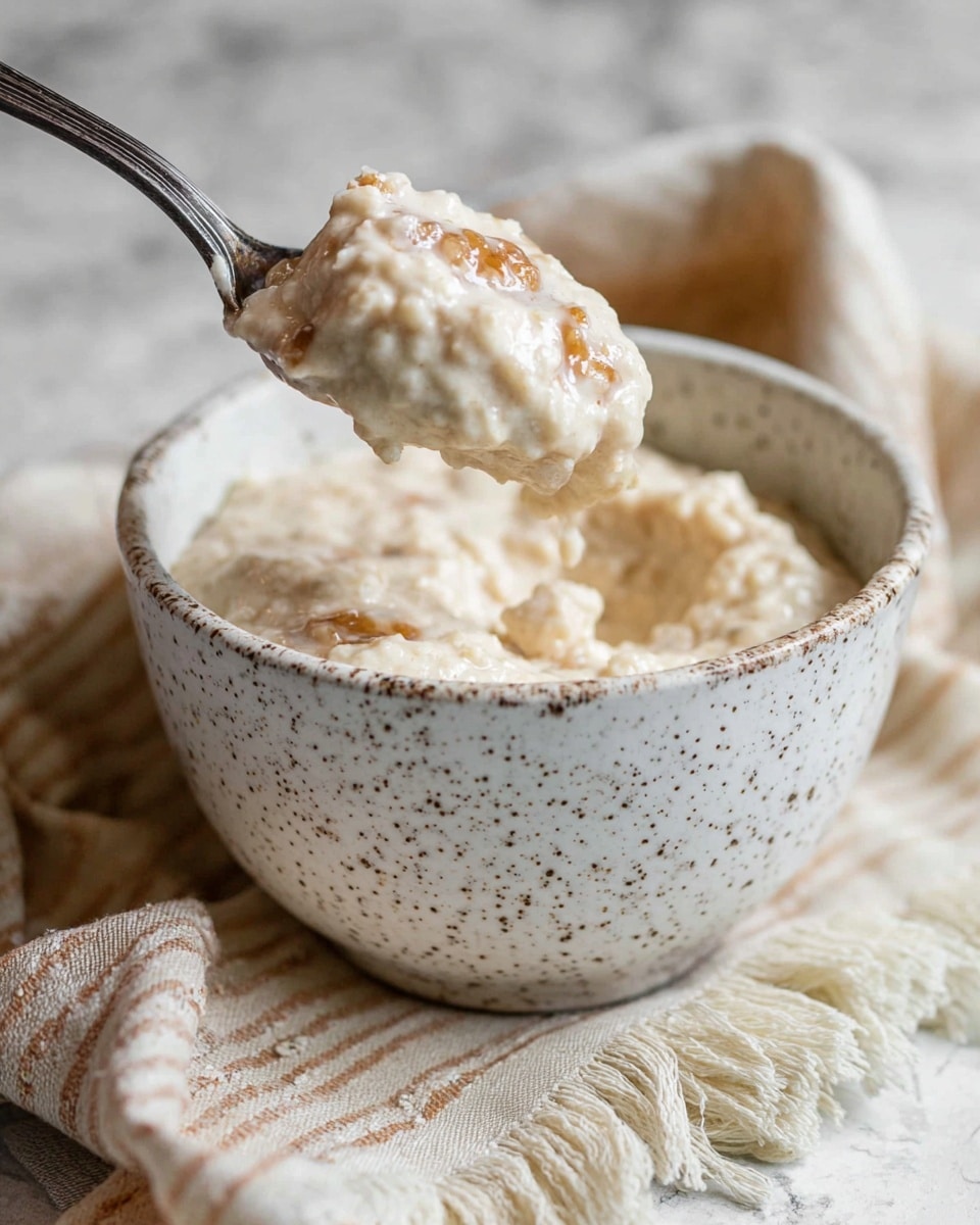 A speckled white bowl filled with a thick, creamy mixture that has a slightly lumpy texture and light beige color, containing small pieces of a golden brown ingredient mixed throughout; a dark metal spoon is lifting a spoonful of the creamy mixture above the bowl, with some mixture hanging off the edge of the spoon. The bowl is placed on a beige and white striped fabric with frayed edges, all set on a white marbled surface. Photo taken with an iphone --ar 4:5 --v 7