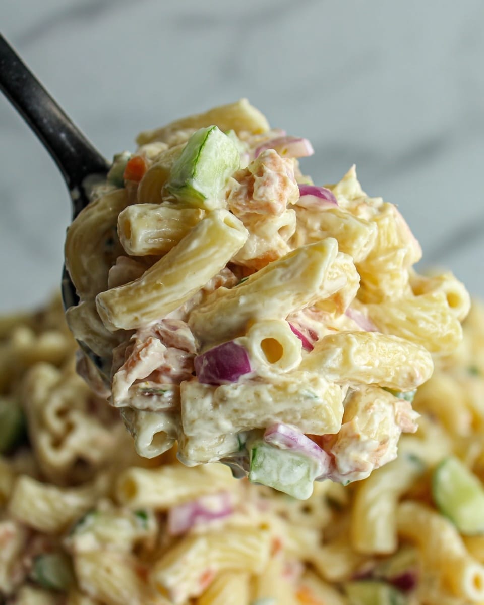 A close-up view of a creamy pasta salad lifted by a black spoon, showing three main layers: small pale yellow pasta tubes as the base, mixed with thin, pale purple shreds and small cubes of light green cucumber; all thoroughly coated in a creamy white dressing with a smooth texture, with some tomato or carrot pieces adding small hints of light orange and pink scattered throughout; the background is a soft white marbled texture. Photo taken with an iphone --ar 4:5 --v 7