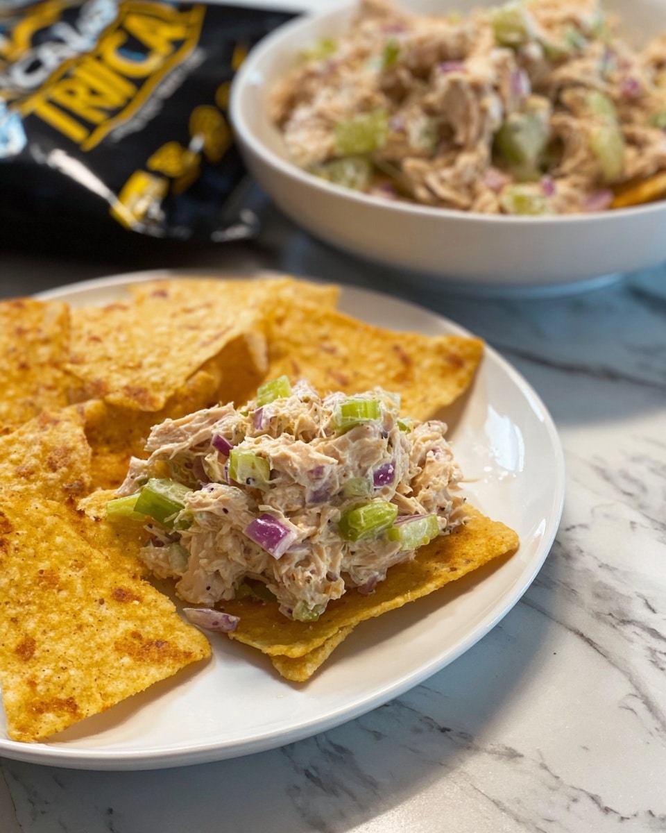 A white plate holds a layered dish with a base of light golden triangular chips topped by a chunky creamy mix made of shredded chicken, green celery pieces, and small red onion cubes, creating a mix of off-white, green, and purple colors with a soft textured look. In the background, a bowl holds more of the chicken mix with the same colors and textures, placed on a white marbled surface. Part of a black snack bag with yellow text is also visible. Photo taken with an iphone --ar 4:5 --v 7