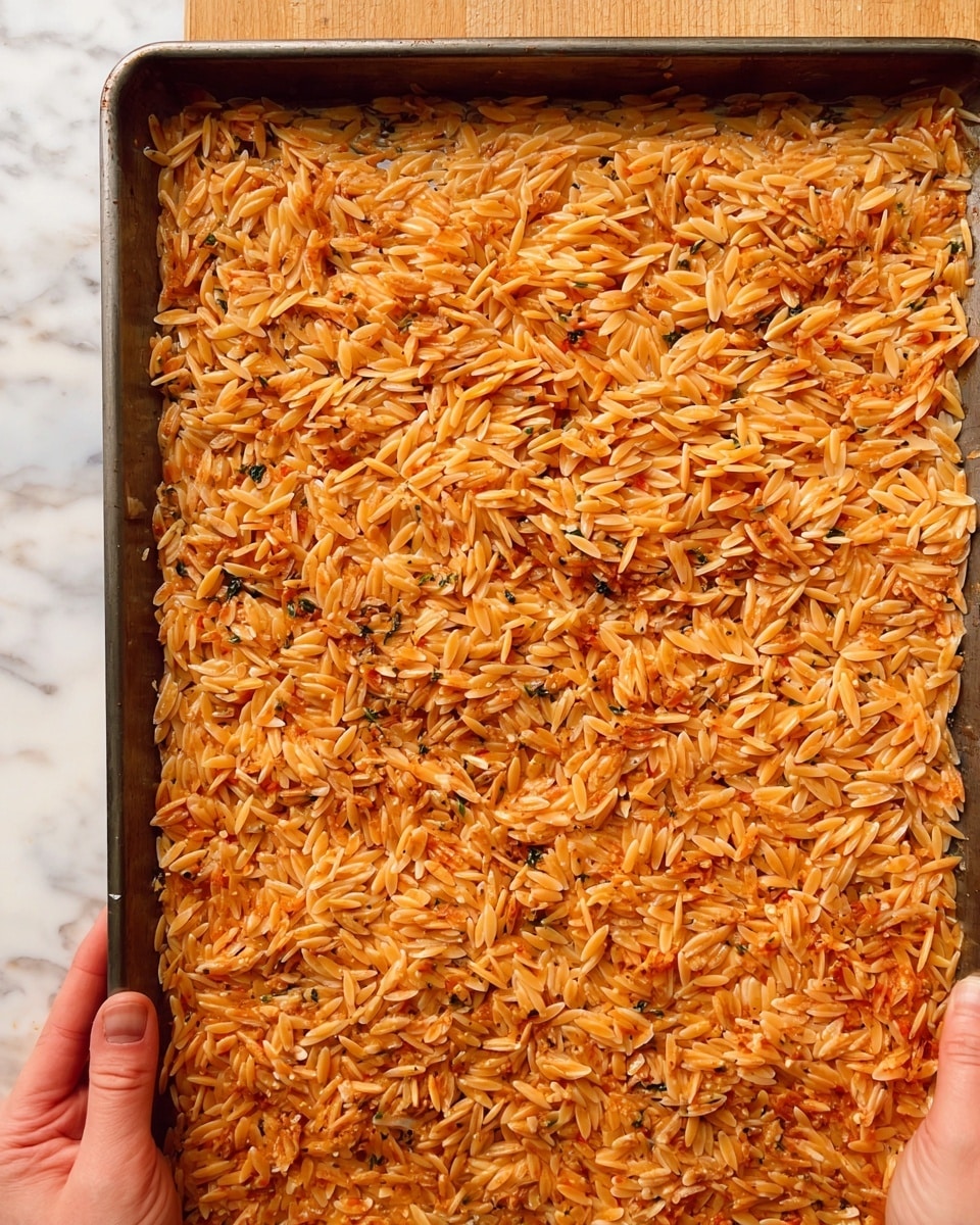 A sheet pan filled with a single layer of cooked orzo pasta, showing a light reddish-orange color mixed throughout. The pasta pieces have a slightly shiny texture and some small bits of seasoning and herbs are scattered evenly over the orzo. Two woman's hands hold the pan from the bottom corners, with a wooden surface beneath and a white marbled texture background. Photo taken with an iphone --ar 4:5 --v 7