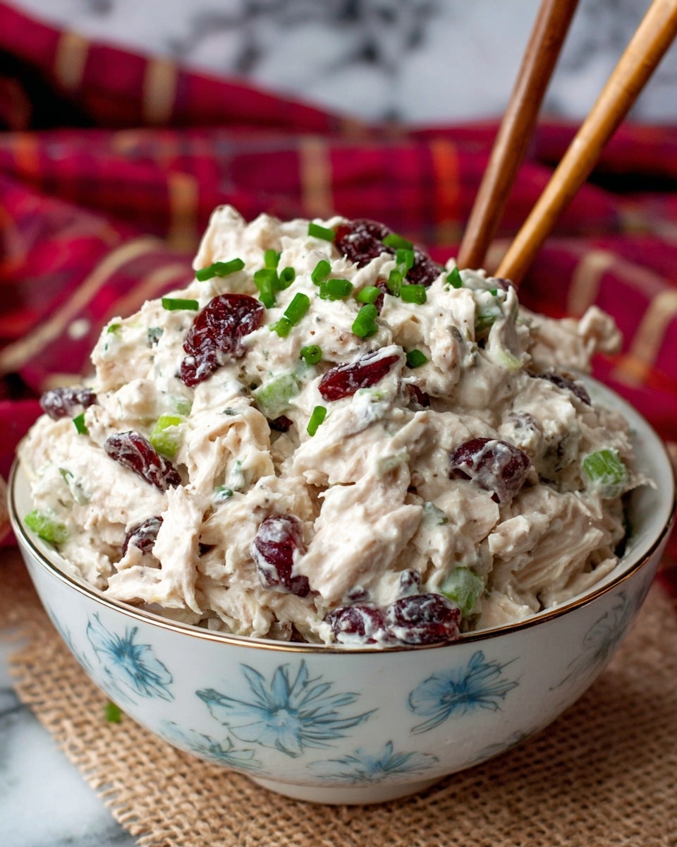 A bowl filled with a chunky, creamy salad made of white shredded chicken mixed with small pieces of green celery and dark red dried cranberries, all coated in a thick white dressing. The salad is garnished with small chopped green chives sprinkled on top. The bowl is white with a subtle blue floral pattern. A pair of wooden chopsticks stands inserted into the salad from the right side. The background shows a soft red plaid cloth, and the bowl sits on a light brown textured mat over a white marbled surface. Photo taken with an iphone --ar 4:5 --v 7
