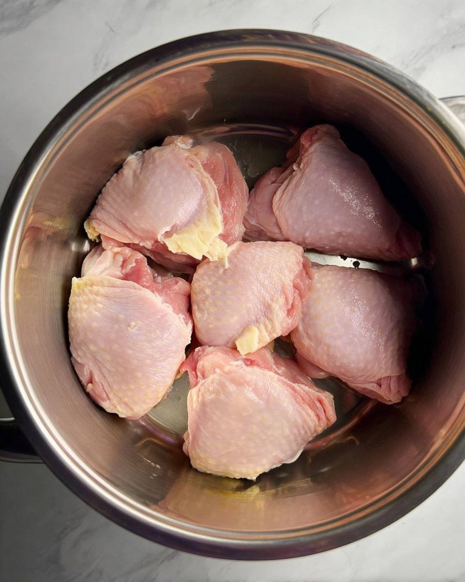 Inside a shiny metal pot, six raw pink chicken pieces are spread out evenly, showing smooth and moist textures with some pale yellow fat still attached. The pot's reflective surface catches the light, adding a slight silver shine to the scene. The background is a white marbled surface visible around the pot's edges. photo taken with an iphone --ar 4:5 --v 7