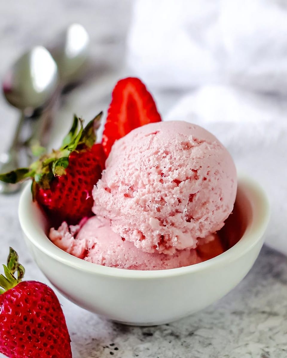 A close-up view of one scoop of light pink strawberry ice cream in a small white bowl, showing a thick and slightly rough texture with tiny air holes on the surface. Behind the scoop, there are fresh red strawberries with green leaves, one whole and one sliced, leaning against the bowl. The bowl sits on a white marbled surface with blurred silver spoons and a white cloth in the background, giving a fresh and simple look. photo taken with an iphone --ar 4:5 --v 7