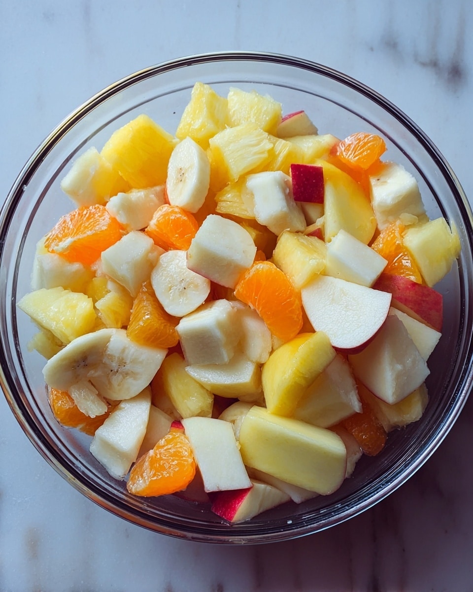 A clear glass bowl filled with a colorful mix of fresh fruit pieces sits on a white marbled surface. The fruit layers include pale yellow pineapple chunks, white banana slices, bright orange mandarin segments, and yellow peach slices with a touch of red skin from apples, all mixed together showing different shapes and smooth textures. The bowl is full but not overflowing, letting each fruit piece show clearly against the glass. Photo taken with an iphone --ar 4:5 --v 7