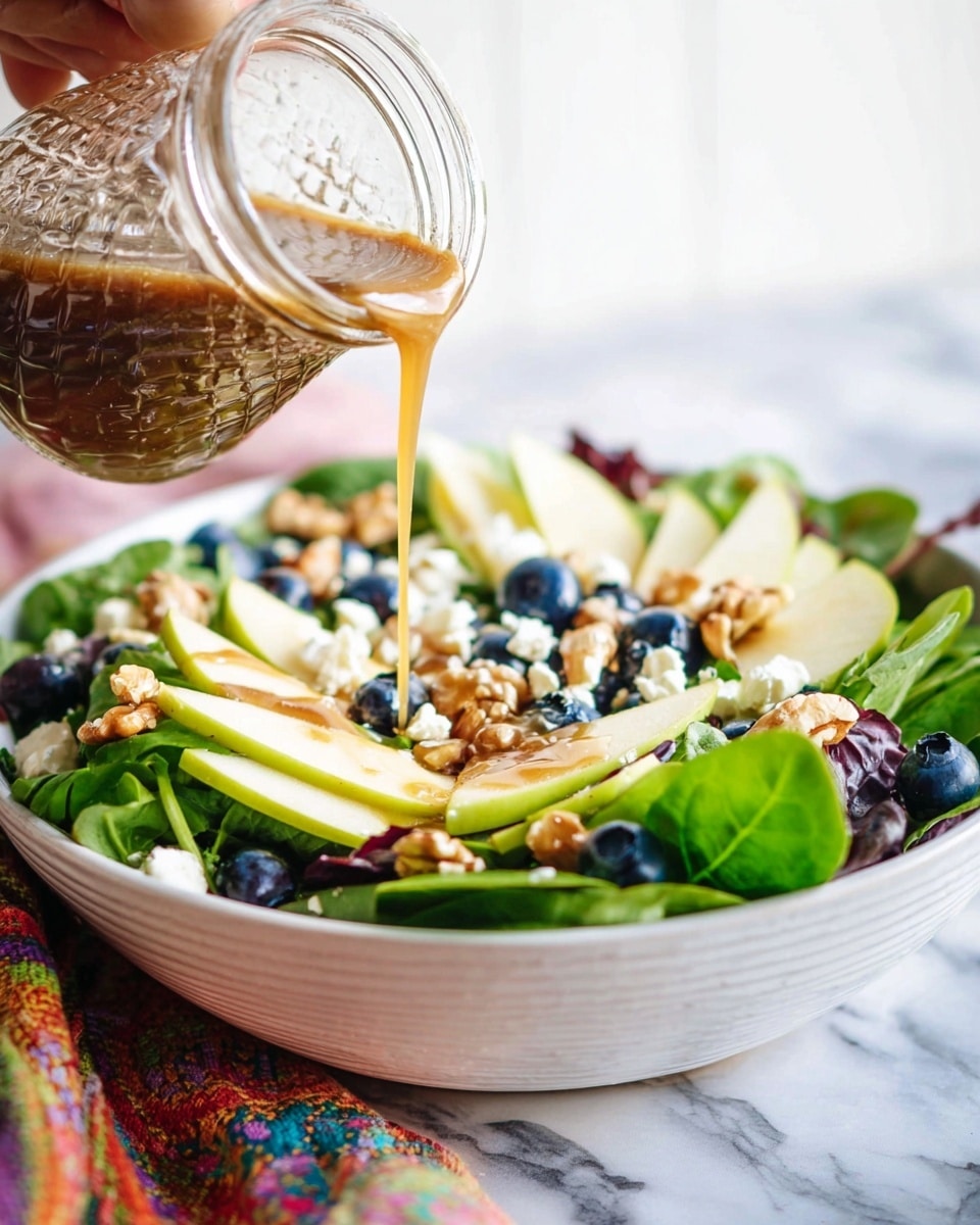 A white bowl filled with a colorful mixed salad layered with dark green leaves at the bottom, topped with thin light yellow apple slices, small dark blue blueberries, white crumbled cheese, and scattered chopped walnuts. A woman's hand is pouring a light brown dressing from a clear textured glass jar over the salad. The bowl is on a white marbled surface with a colorful cloth partially visible underneath. photo taken with an iphone --ar 4:5 --v 7
