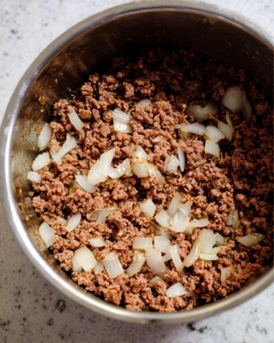 A close-up of a silver pot filled with cooked ground meat mixed with small pieces of translucent white onion. The meat is brown and crumbly, spread evenly throughout the pot, while the onion pieces add texture and light color contrast. The pot sits on a white marbled surface, reflecting a warm light. photo taken with an iphone --ar 4:5 --v 7