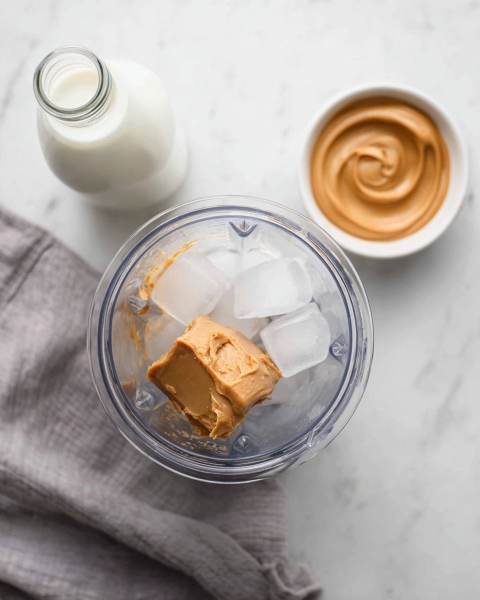 The image shows a top view of a clear blender cup with ice cubes, a large scoop of light brown peanut butter, and a splash of creamy white liquid inside. Behind the blender, there is a glass bottle filled with white milk on the left and a small white bowl with a smooth swirl of peanut butter on the right. The setup is on a white marbled surface with a soft grey cloth folded to the left side. Photo taken with an iphone --ar 4:5 --v 7