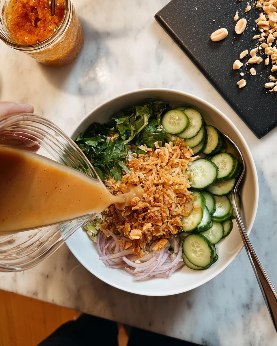 A white bowl holds a layered dish with four sections: thinly sliced cucumber with dark green edges arranged neatly at the top right, thin light pink onion slices next to the cucumbers, a cluster of fresh dark green herbs below the onions, and a generous portion of crispy golden-brown fried rice piled in the center. A silver spoon rests inside the bowl along the left edge. A woman's hand is pouring a light brown sauce from a clear ribbed jar towards the bowl. In the background, a black cutting board with scattered chopped peanuts rests on a white marbled surface, with a dark container of orange food visible at the top right. photo taken with an iphone --ar 4:5 --v 7