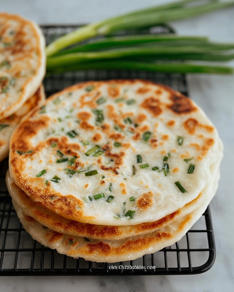 The image shows three round, lightly browned flatbreads stacked slightly overlapping on a black wire rack. Each flatbread has a golden-brown crispy texture with small bubbles and darker brown spots scattered on the surface. Green chive pieces are embedded throughout the white dough, adding small bursts of green color. In the background, some fresh green onions are placed next to the flatbreads. The whole scene is set on a white marbled surface. photo taken with an iphone --ar 4:5 --v 7