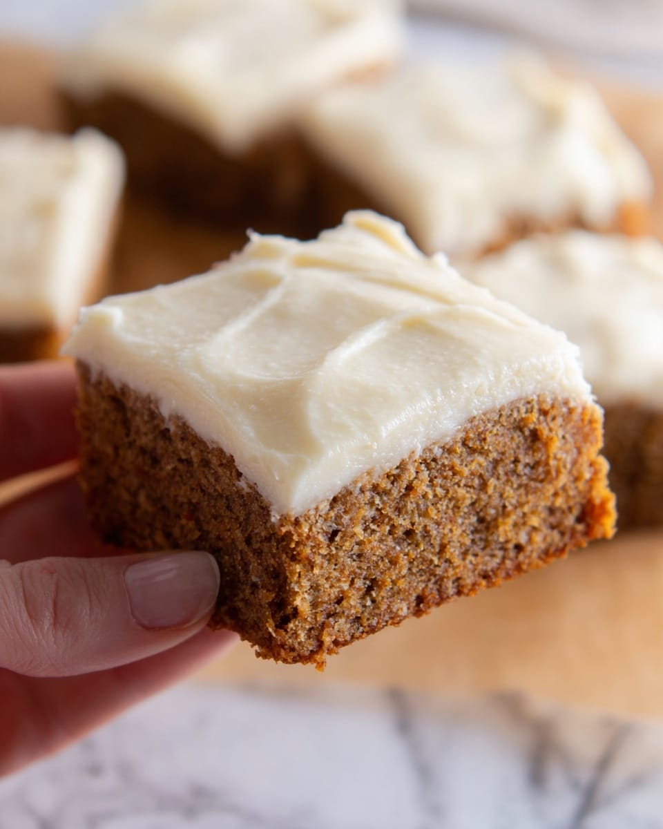 The image shows a close-up of a single square piece of cake held by a woman's hand. It has two layers: the bottom layer is a moist, brown cake with a slightly crumbly texture, and the top layer is a thick, creamy, smooth, and pale white frosting that looks soft and spread unevenly but nicely across the cake. The background features other similar cake pieces blurred out on a white marbled surface. Photo taken with an iphone --ar 4:5 --v 7