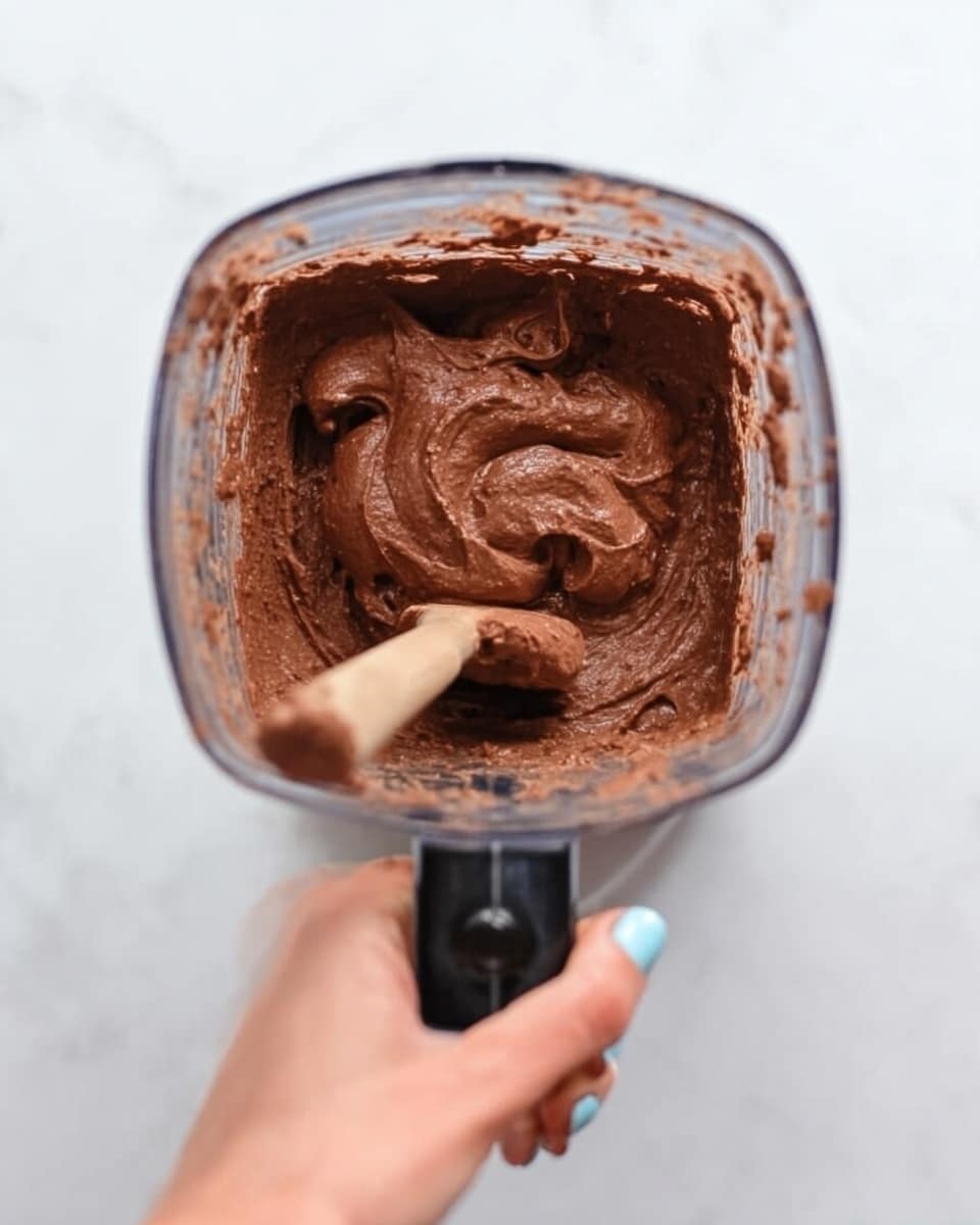 The image shows a clear blender container filled with thick, creamy chocolate mixture with a smooth and dense texture. A light brown wooden spoon sticks out from the mixture, angled slightly towards the viewer. A woman's hand with light skin tone and pale blue nail polish holds the black blender base from the bottom left corner. The background is a white marbled texture, giving a clean, bright look to the scene. photo taken with an iphone --ar 4:5 --v 7