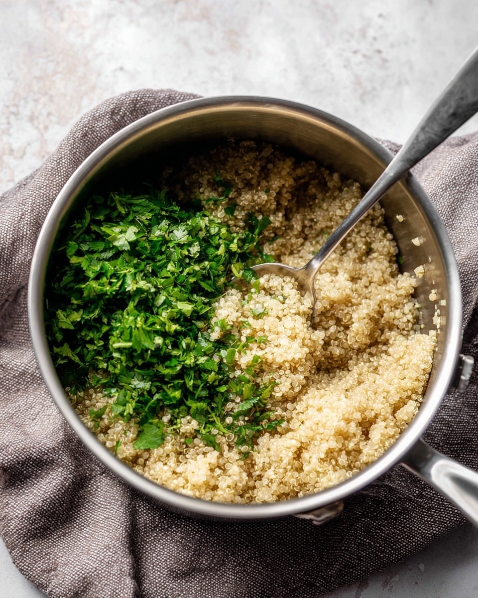 Inside a silver cooking pot, there is a mix of light beige cooked quinoa covering most of the bottom and right side. On top and left side, there is a pile of fresh chopped green herbs with leafy textures scattered unevenly. A silver spoon is partly inside the pot on the right, scooping up some quinoa, with the handle pointing outward. The pot sits on a textured gray cloth, all set on a white marbled surface. photo taken with an iphone --ar 4:5 --v 7