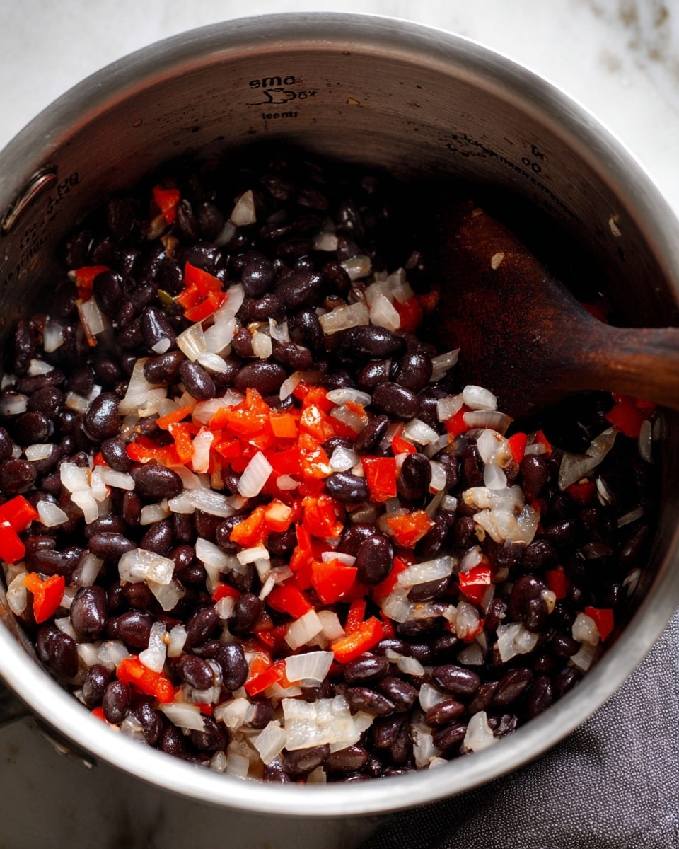 A close-up view inside a silver metal pot showing a mix of cooked black beans, chopped white onions, and red bell pepper pieces. The black beans have a shiny texture, scattered evenly with the translucent diced onions and bright red pepper chunks. A dark wooden spoon partially rests in the mixture on the right side. The pot has measurement markings on the inside near the top edge. A gray cloth is partly visible under the pot on a white marbled surface. Photo taken with an iphone --ar 4:5 --v 7