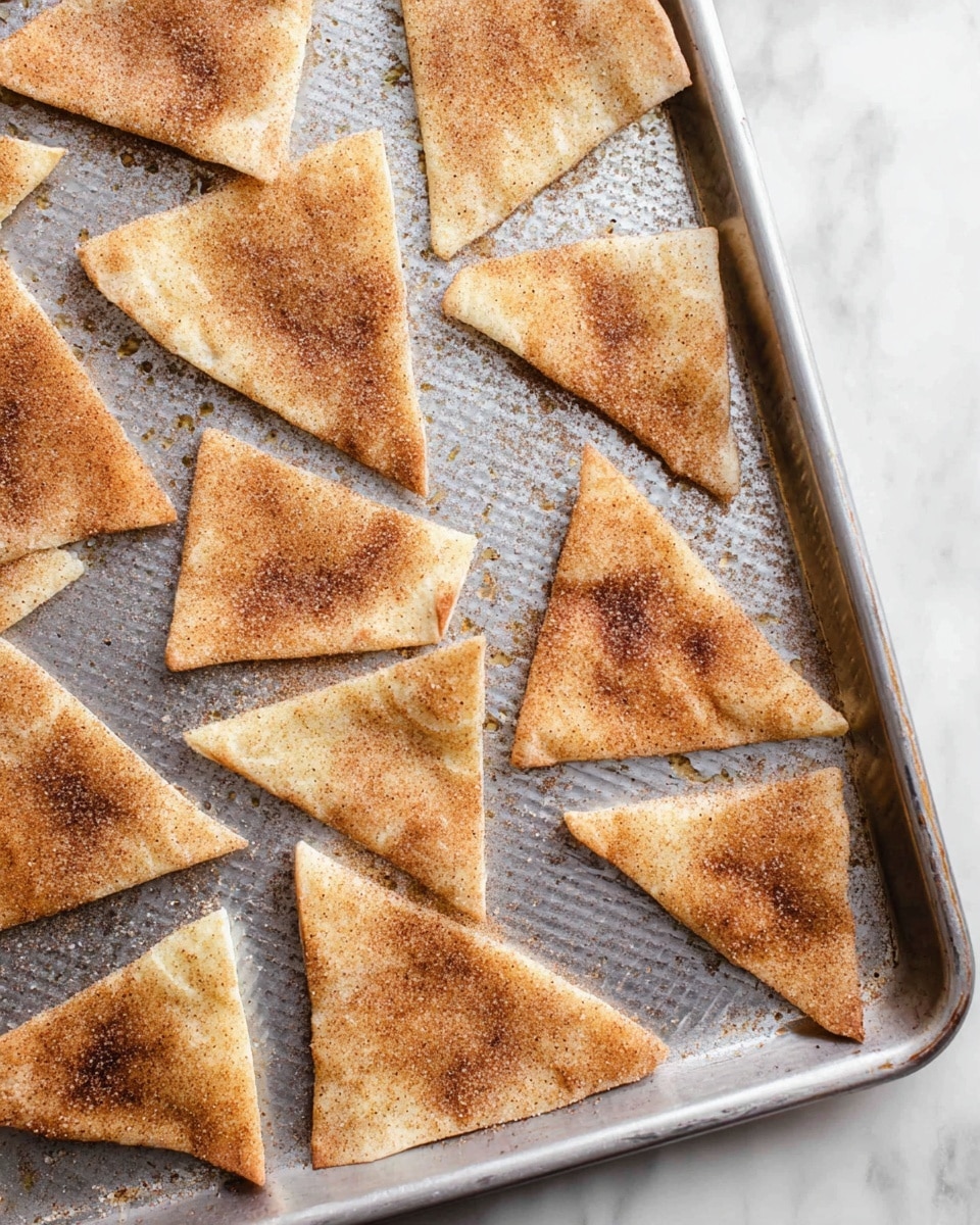 Triangles of toasted pita bread are arranged in a single layer on a metal baking sheet with a textured surface. Each pita piece is light golden brown with a visible sprinkle of cinnamon and sugar, giving a light grainy texture on top. The pita triangles vary slightly in size and each piece shows small toasted spots, adding a slightly uneven color. The metal baking sheet has a slightly shiny surface with small drops of oil around some edges of the pita pieces. The whole scene sits on a white marbled surface. photo taken with an iphone --ar 4:5 --v 7