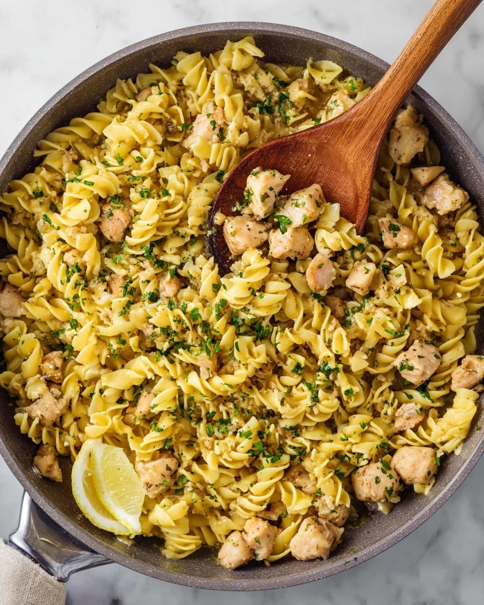 In a grey pan on a white marbled surface, there is a dish with three main layers: the bottom layer is a mix of small chicken pieces, light brown with a soft texture, scattered evenly throughout; the middle layer is pasta with a unique yellow color and ruffled, wavy shapes, filling the pan fully and mixed with the chicken; the top layer has small green herb pieces sprinkled all over for color and freshness. A wooden spoon is scooping out a portion showing all layers together, and a small slice of lemon is resting near the pan’s edge. Photo taken with an iphone --ar 4:5 --v 7