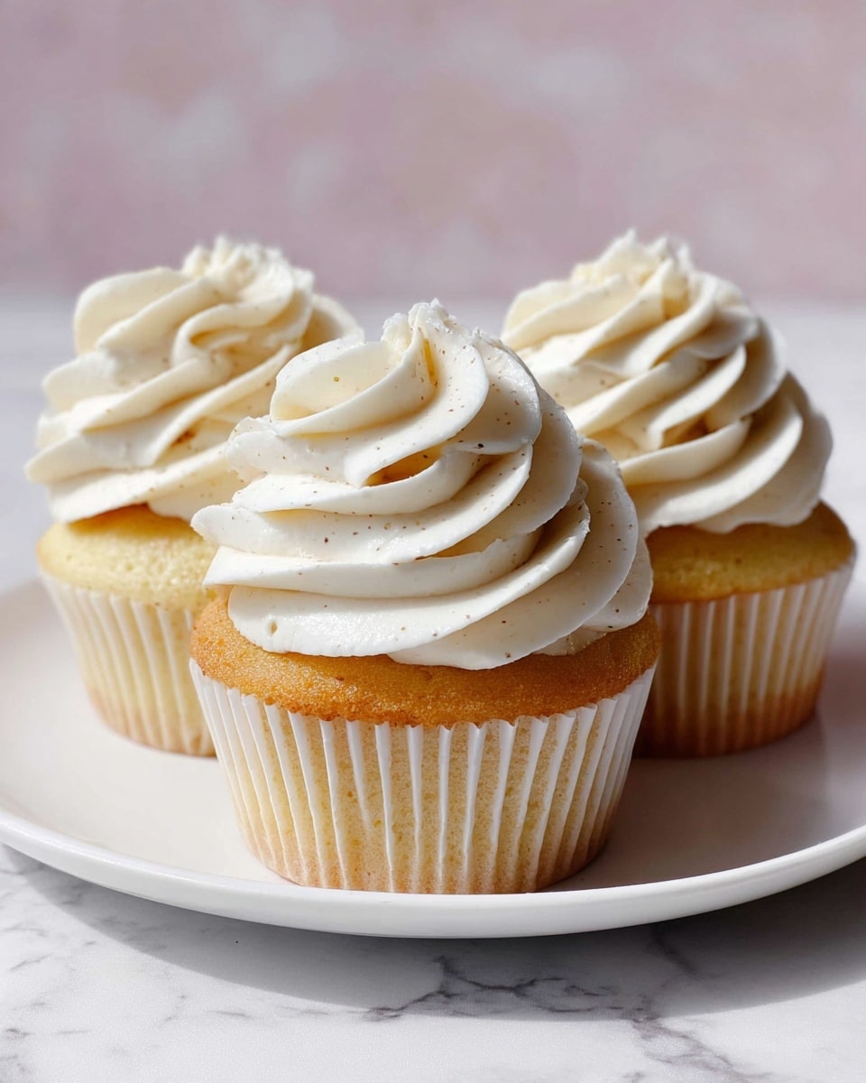Three vanilla cupcakes sit closely together on a white plate resting on a white marbled surface. Each cupcake has one thick swirl of creamy white frosting on top, showing smooth textures and gentle peaks, with tiny specks of vanilla visible throughout. The golden yellow cake bases peek through the plain white cupcake liners. The overall look is soft and inviting with a slight shine on the frosting. photo taken with an iphone --ar 4:5 --v 7