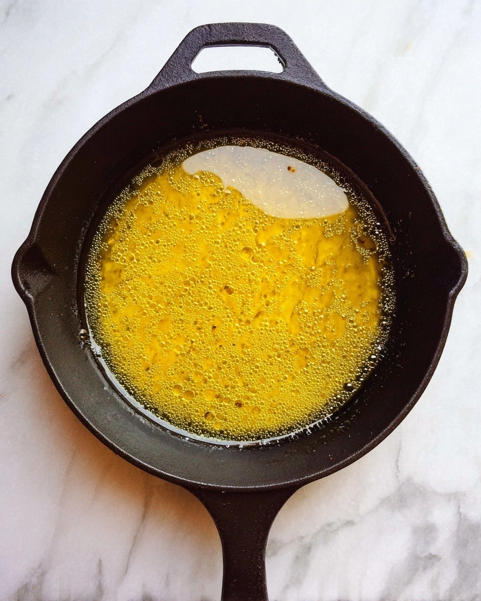 A dark cast iron pan filled with melted golden yellow butter, showing small bubbles and patches of slightly darker brown spots on the surface, resting on a white marbled texture. The pan is centered, viewed from above, with a handle extending downward and a small handle on the opposite side, both in cast iron texture photo taken with an iphone --ar 4:5 --v 7