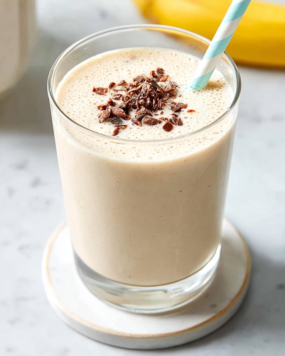 A clear glass filled with a creamy, pale beige smoothie that has a smooth, slightly frothy texture on top. The smoothie is topped with small dark brown clusters that add texture and contrast. A light blue and white striped straw is placed in the smoothie on the right side. The glass sits on a round white coaster, all set on a surface with a white marbled texture. In the blurred background, a yellow banana is visible on the right side, adding a fresh touch. Photo taken with an iphone --ar 4:5 --v 7