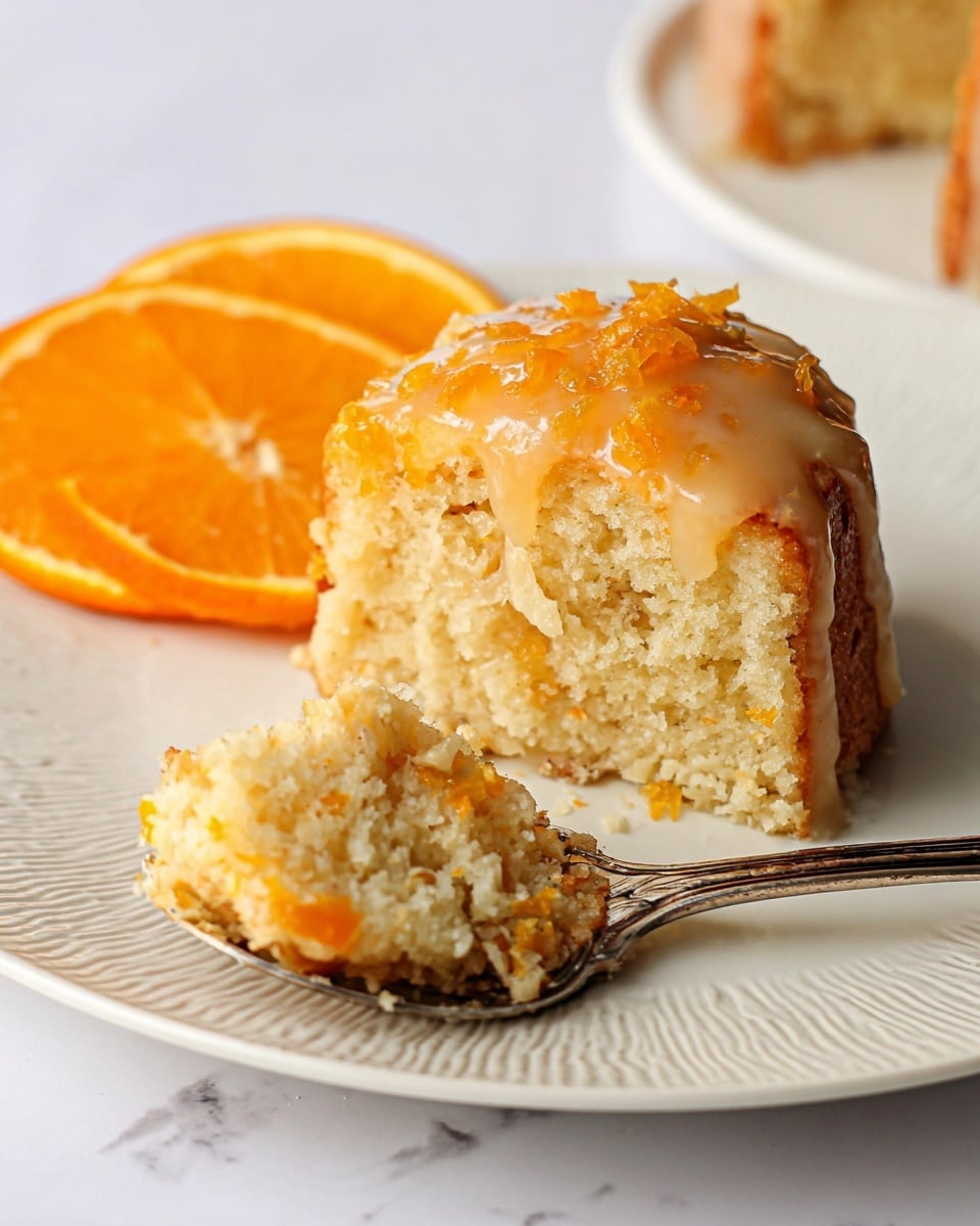 A close-up image of a soft, light beige cake with bits of orange peel mixed inside, topped with a glossy orange glaze. The cake is shown with a small piece on a silver spoon in the foreground, while the main piece has a warm orange shine on top. Behind the cake are two fresh orange slices adding a bright orange color. All are placed on a white plate with a textured rim, set on a white marbled surface. photo taken with an iphone --ar 4:5 --v 7