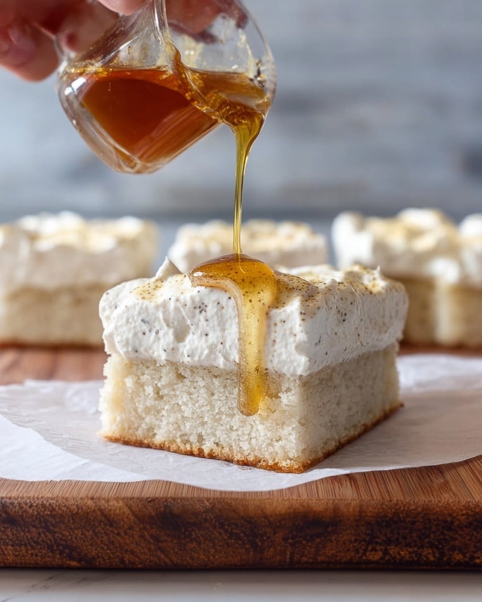 The image shows a close-up of a square dessert with two layers placed on a white parchment paper on a wooden surface. The bottom layer is a pale, soft, and moist cake with a slightly grainy texture. The top layer is a thick, creamy, white frosting with small visible specks, giving it a textured look. A woman's hand is pouring golden brown syrup with visible tiny seeds or spices onto the top layer, with the syrup flowing down the side. In the background, there is a blurred view of more pieces of the same dessert on a white marbled surface. photo taken with an iphone --ar 4:5 --v 7